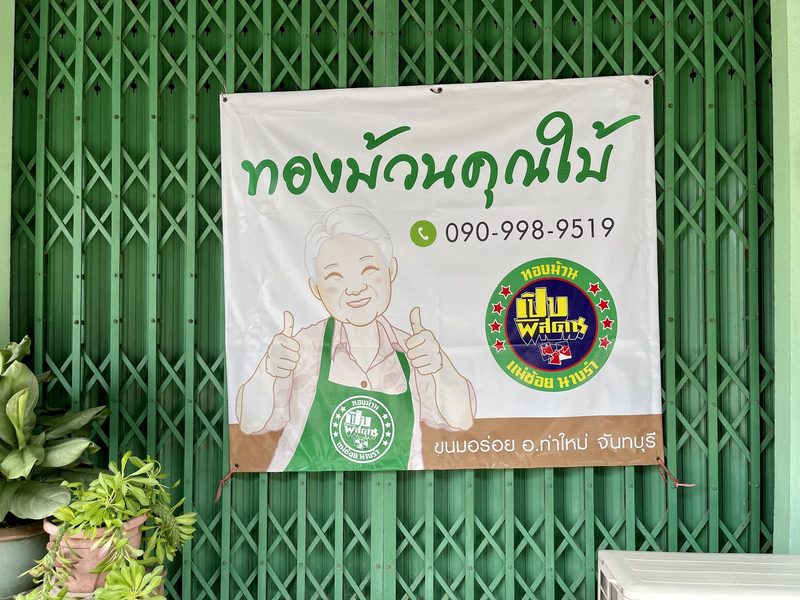 ป้ายผ้าชื่อร้านทองม้วนคุณใบ้ มีรูปการ์ตูนคุณยายยกนิ้วโป้ง แขวนบนประตูเหล็กยืดสีเขียว
