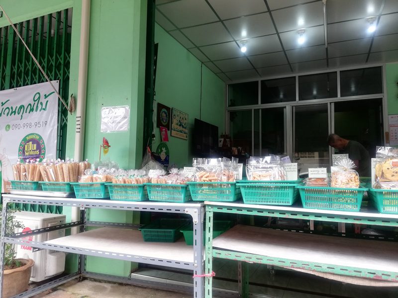 ในร้านมีโต๊ะยาววางตะกร้าสีเขียวใส่ขนมบรรจุถุงเรียงเต็มแถว