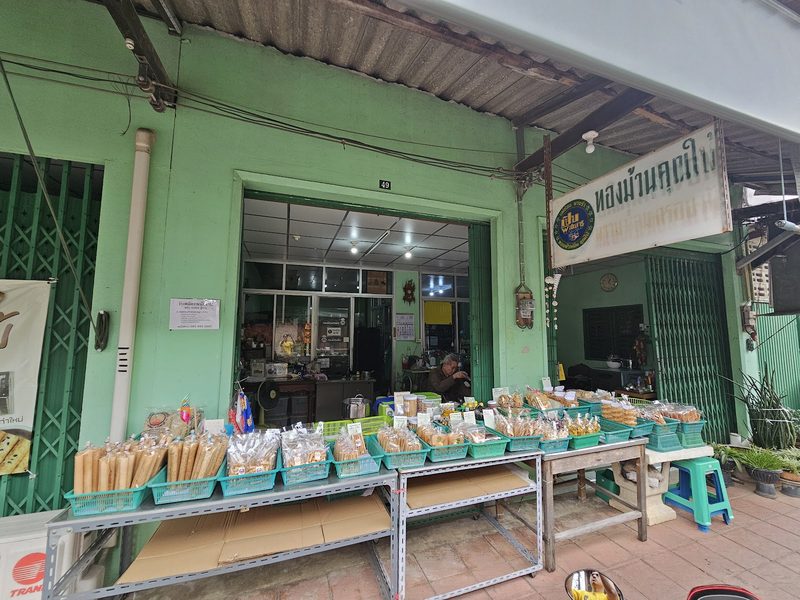 หน้าร้านทองม้วนคุณใบ้ ห้องแถวสีเขียวมีป้ายชื่อร้าน และโต๊ะวางตะกร้าขนมเรียงยาวหน้าร้าน
