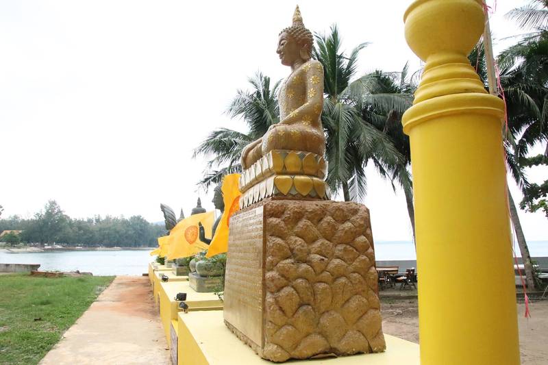 พระพุทธรูปสีทองบนแท่นริมทางเดิน มีทะเลและต้นมะพร้าวอยู่ด้านหลัง